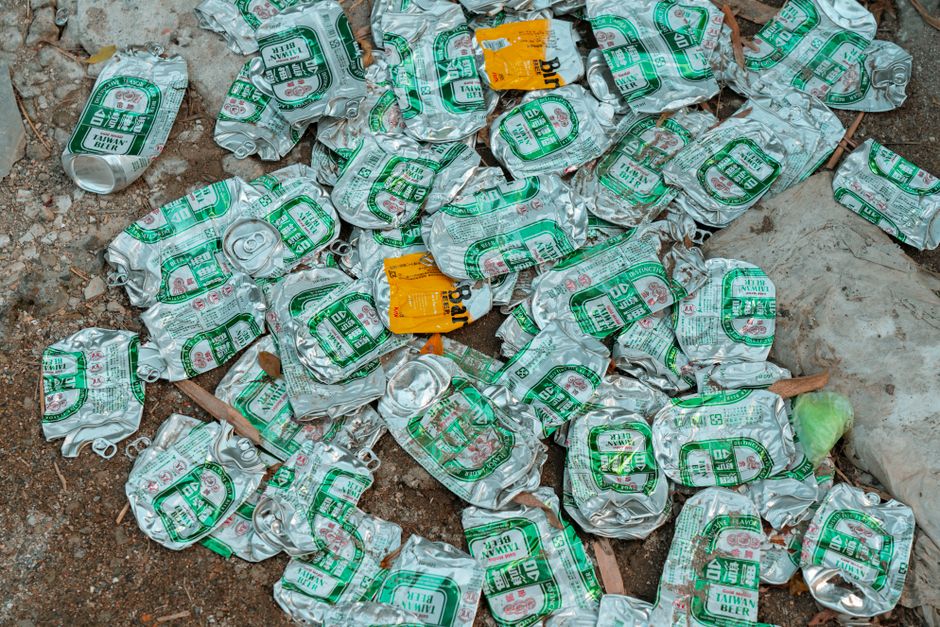 A pile of mixed waste materials is seen on the ground against a rough, light-colored brick wall, adjacent to a darker brick vertical surface and a tree trunk on the left. The rubbish includes flattene