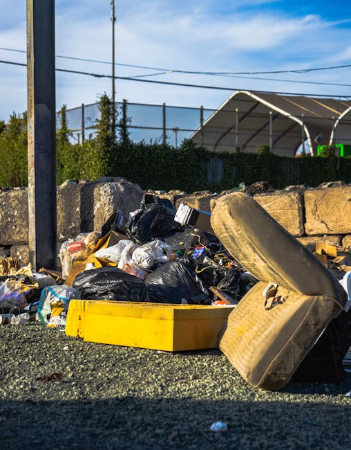 A black wheeled rubbish bin positioned on the pavement beside a residential street at night. The bin, labeled with 'ST. JOHN'S', contains a variety of dumped waste, including cardboard boxes and other